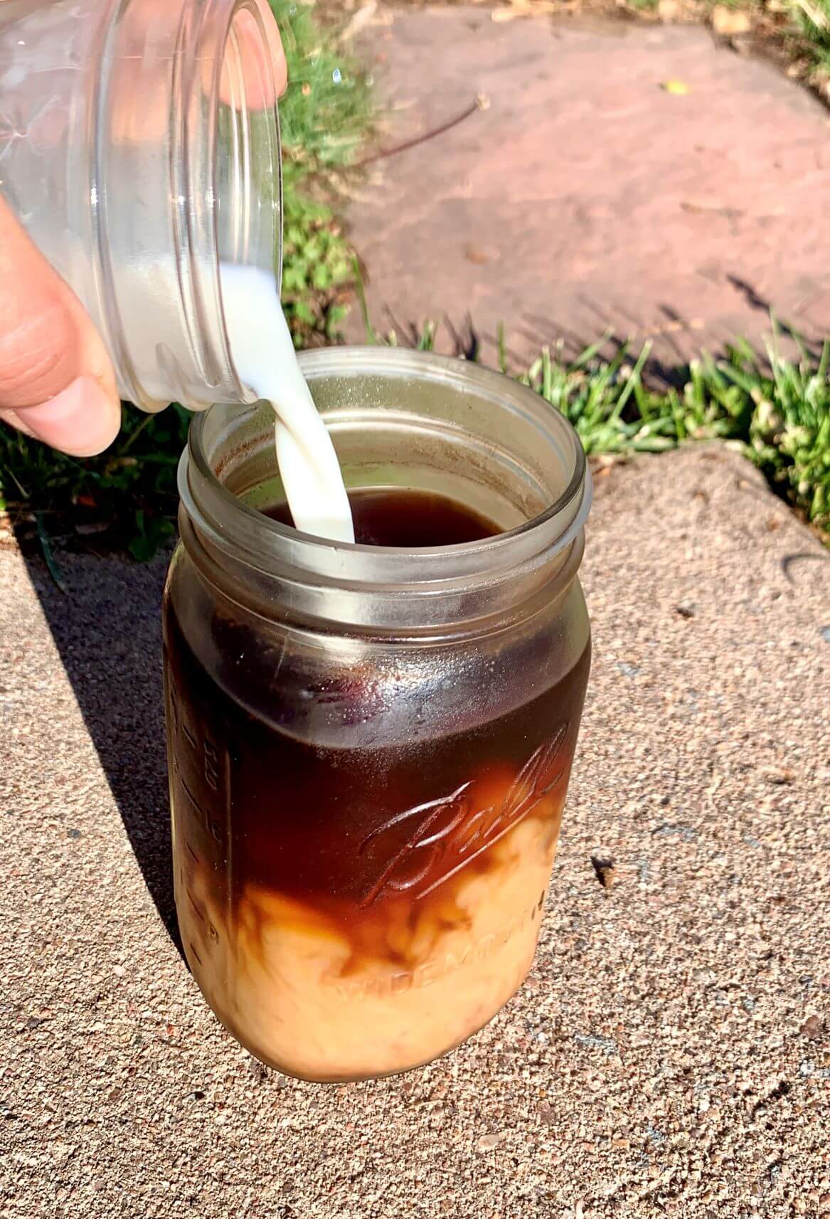 iced coffee in a mason jar with milk being poured in from small mason jar, on concrete with flagstone and grass behind it.