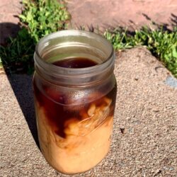 iced coffee in a mason jar with milk poured in, all on concrete with flagstone and grass behind it.