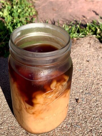 iced coffee in a mason jar with milk poured in, all on concrete with flagstone and grass behind it.