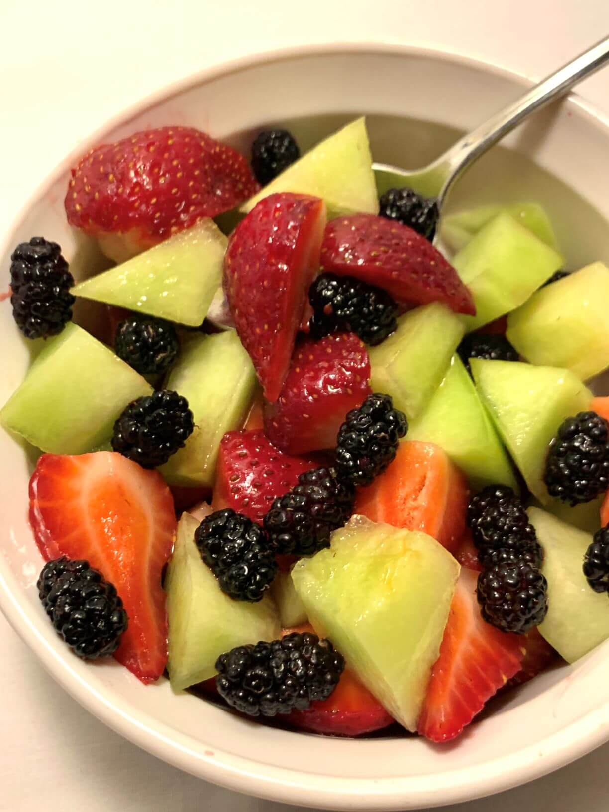 fruit in white corning ware bowl with stainless steel spoon. Fruit consists of chopped lemon drop melon, strawberries, and mulberries.