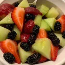 fruit in white corning ware bowl with stainless steel spoon. Fruit consists of chopped lemon drop melon, strawberries, and mulberries.