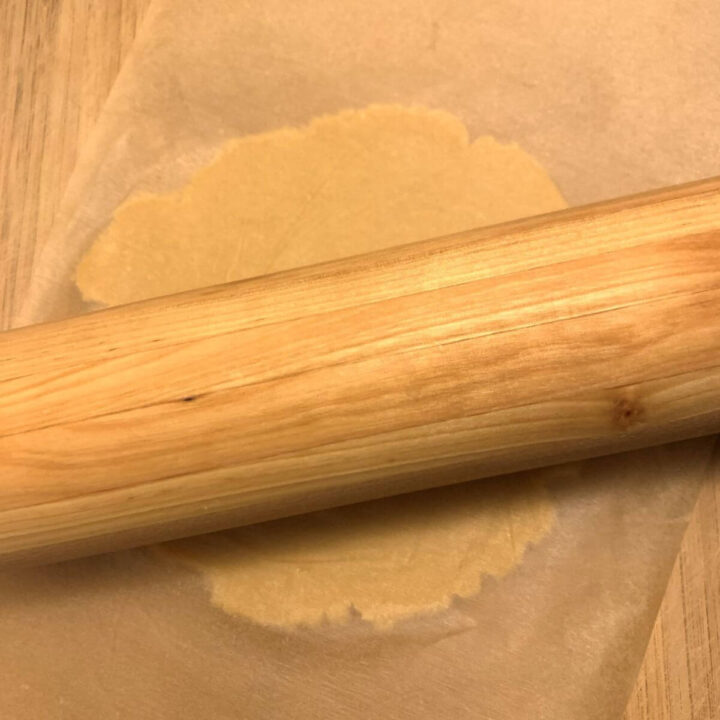 cookie dough being rolled out between 2 pieces of parchment paper with a wooden rolling pin.