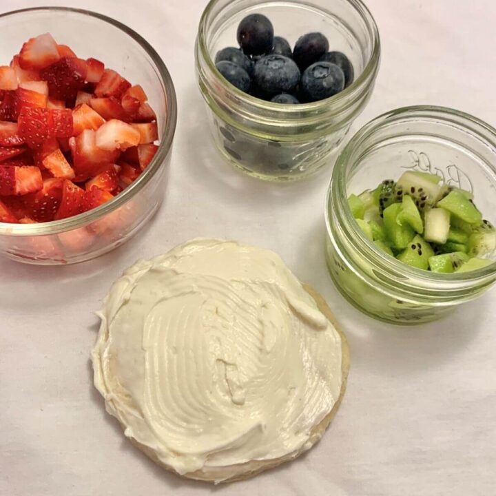 1 sugar cookie with creamy white frosting surrounded by diced strawberries, kiwi, and blueberries in small glass jars, all on a white towel.