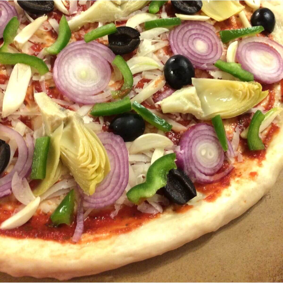 pizza dough with sauce, shredded cheese, garlic, olives, green bell pepper, boiler onions, artichoke hearts, all on a pizza stone.