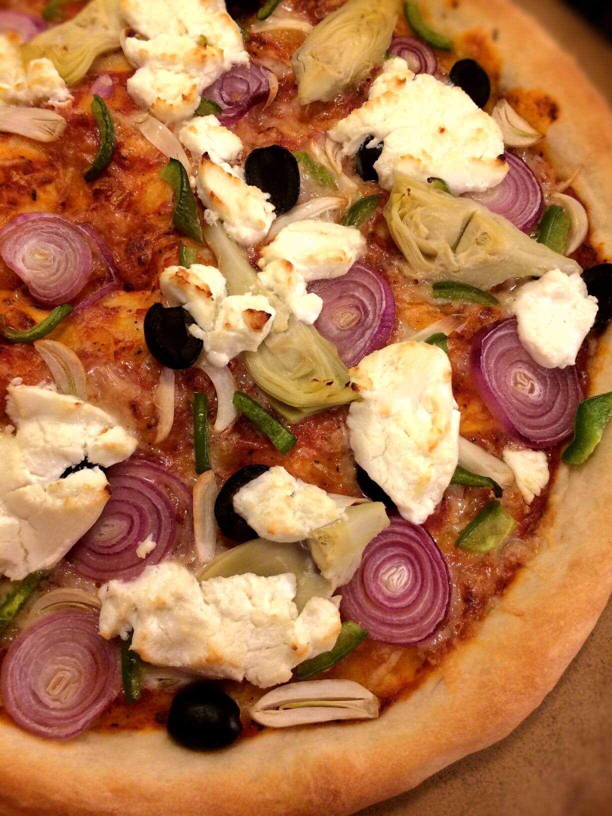 Baked pizza with sauce, shredded cheese, garlic, olives, green bell pepper, boiler onions, artichoke hearts, and goat cheese clumps all on a pizza stone.