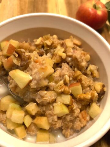 Square image of apple pie oatmeal with silver spoon scooping diced apples, walnuts and oatmeal in milk in a white CorningWare bowl on wood cutting board with a small apples and walnut pieces running along right side of image.