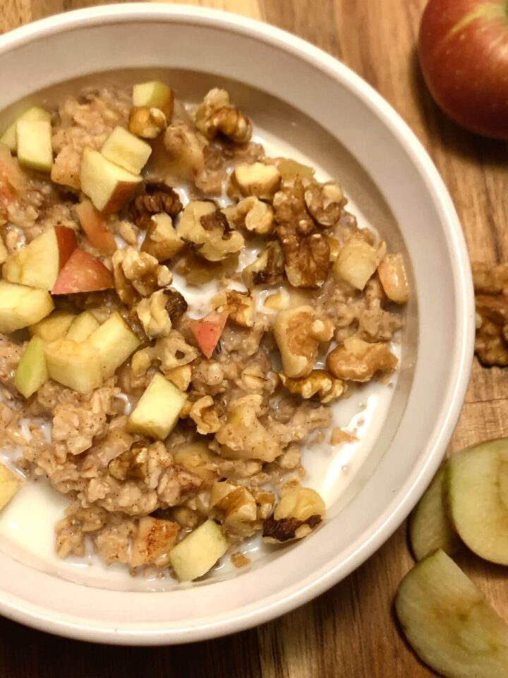 Vertical image of apple pie oatmeal with diced apples, walnuts and oatmeal in milk in a white CorningWare bowl on wood cutting board with a small apple, walnut pieces, and sliced apples running along right side of image.
