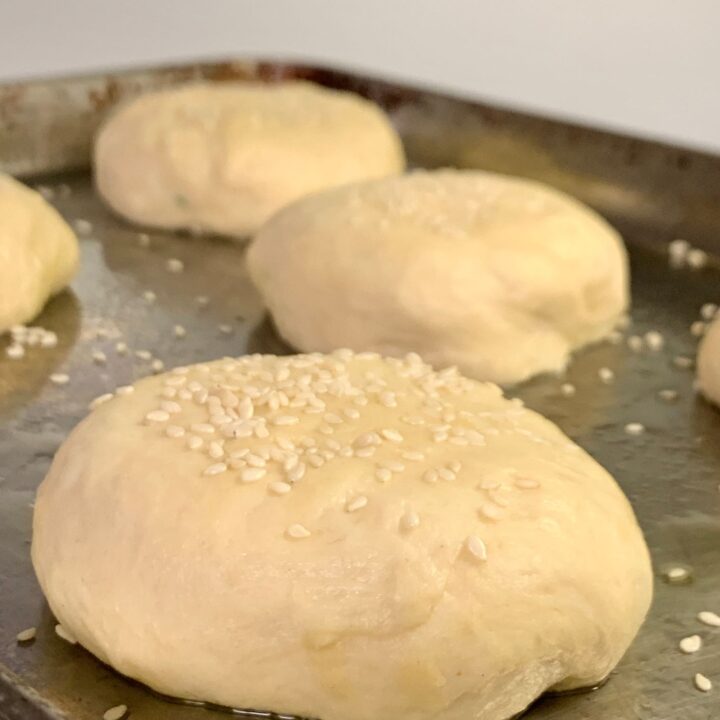 uncooked burger buns portioned out on stainless steel baking sheet with sesame seeds on top.