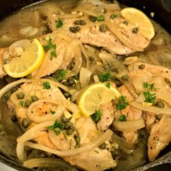 Square image of cooked chicken piccata with chicken breasts, sliced onion, capers, garlic, lemon slices, and parsley in a juice all in cast iron skillet.
