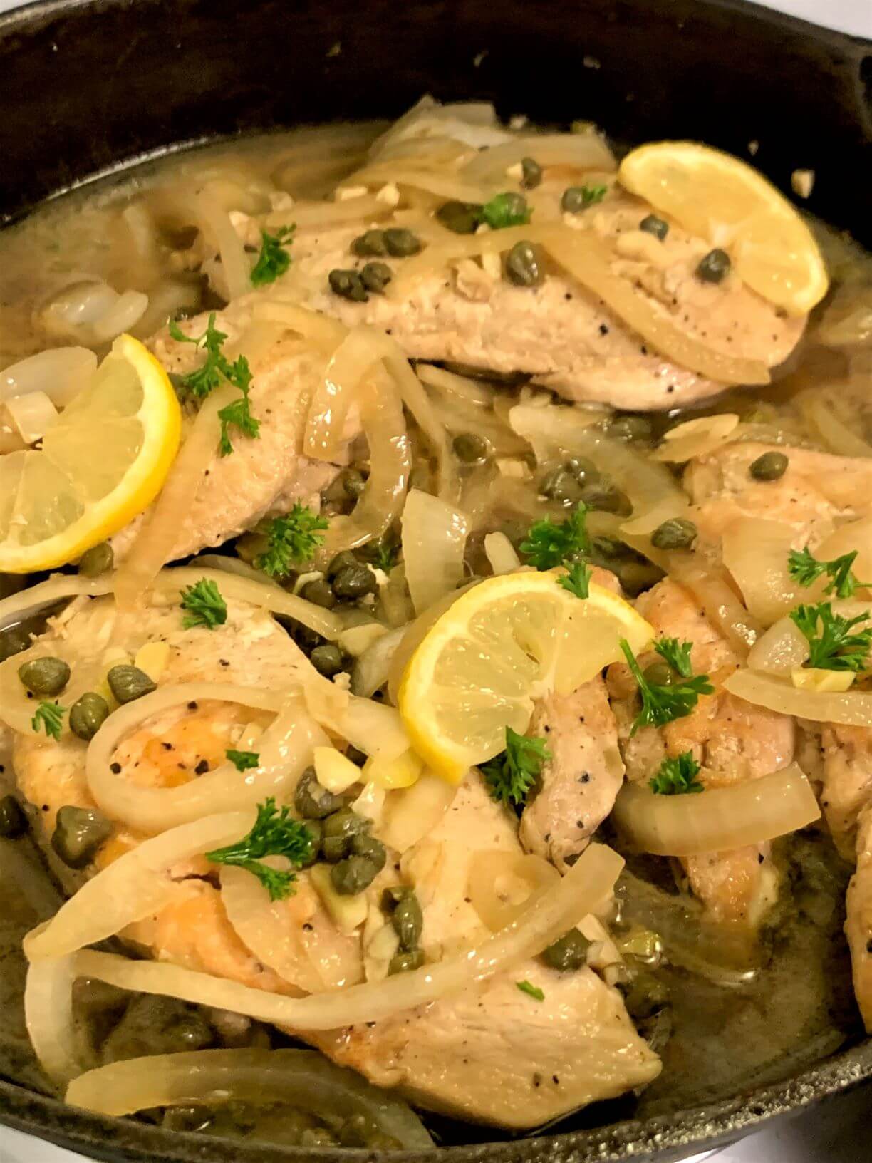 Vertical image of cooked chicken piccata with chicken breasts, sliced onion, capers, garlic, lemon slices, and parsley in a juice all in cast iron skillet.