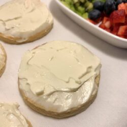 cut out sugar cookies frosted with cream cheese frosting with a white plate of diced fruit and berries partially visible in upper right corner, all on a white tea cloth.