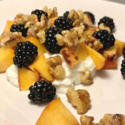 Square image of white oval CorningWare plate with Greek yogurt, diced peaches, blackberries, and walnut pieces covered in honey and vanilla extract.