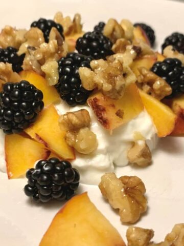 Square image of white oval CorningWare plate with Greek yogurt, diced peaches, blackberries, and walnut pieces covered in honey and vanilla extract.