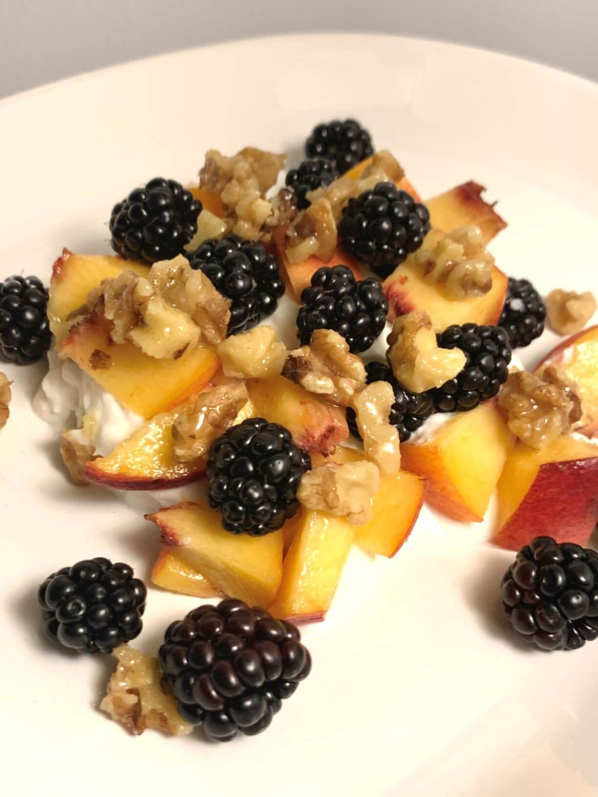 Vertical image of white oval CorningWare plate with Greek yogurt, diced peaches, blackberries, and walnut pieces covered in honey and vanilla extract