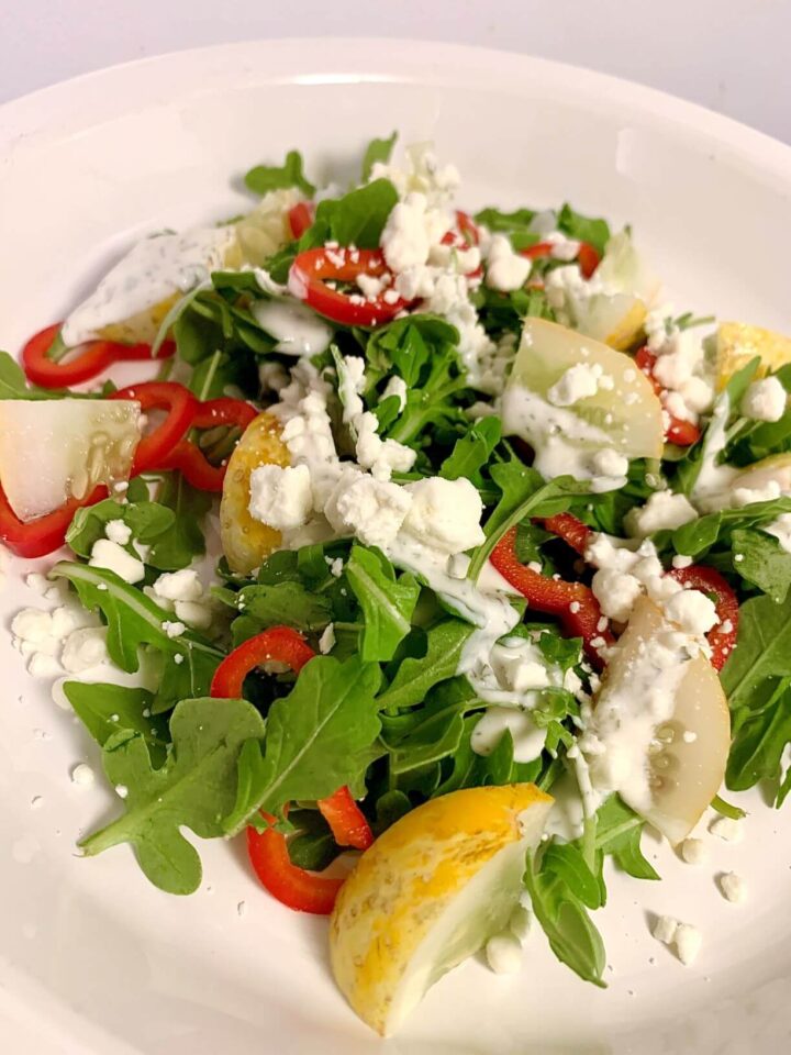 Vertical image of arugula salad with chopped lemon cucumber, sliced mini red bell peppers, goat cheese crumbles, ranch dressing on top, all on white round plate.