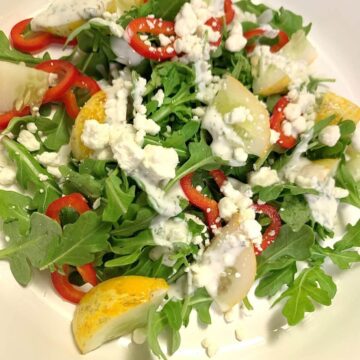 Square image of arugula salad with chopped lemon cucumber, sliced mini red bell peppers, goat cheese crumbles, ranch dressing on top, all on white round plate.