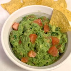 Square image of guacamole with diced tomatoes on top in a Corning Ware bowl with yellow chips, all on white cloth