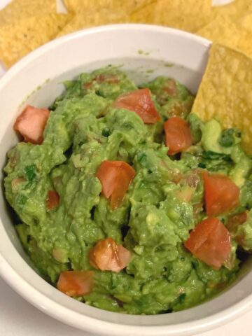 Square image of guacamole with diced tomatoes on top in a Corning Ware bowl with yellow chips, all on white cloth