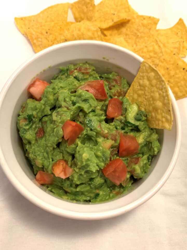 guacamole with diced tomatoes on top in a Corning Ware bowl with yellow chips, all on white cloth